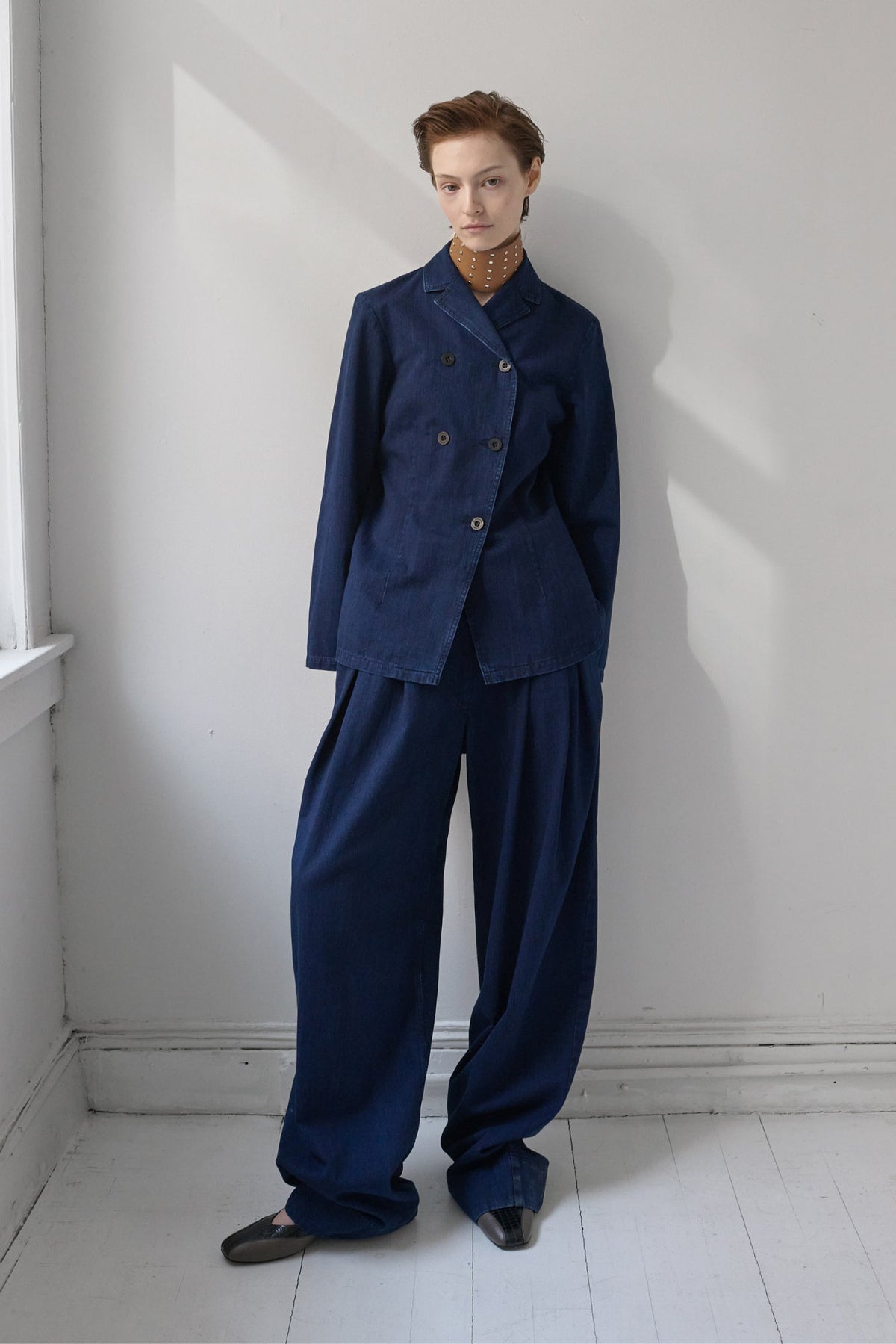 A model stands in a minimalist white room wearing a dark navy blue two-piece suit consisting of a double-breasted jacket and wide-leg trousers, layered over a brown necklace